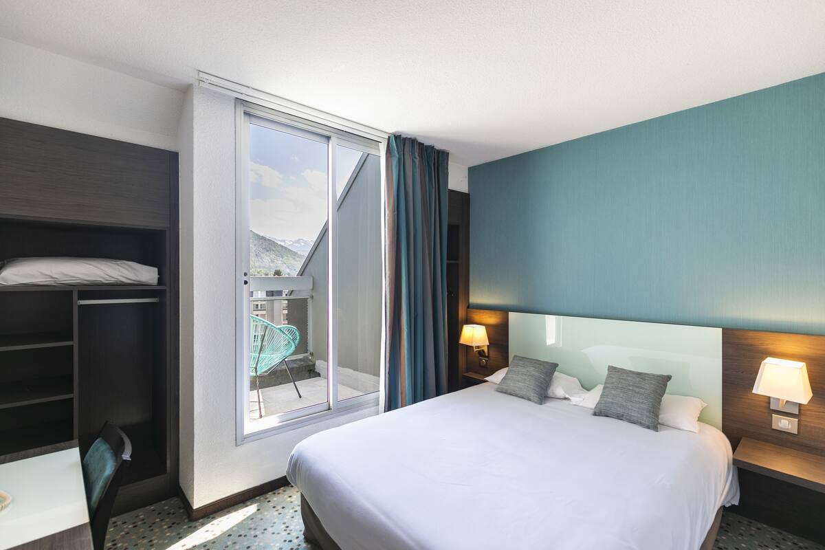 A hotel room with a large bed, a desk, a chair, a wardrobe, a balcony with a chair, and a view of the mountains.