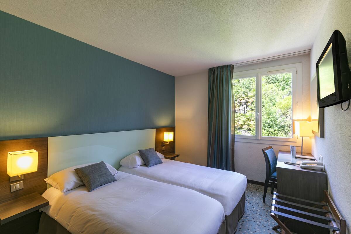 A hotel room with two beds, a desk, a chair, a TV, and a window with curtains.