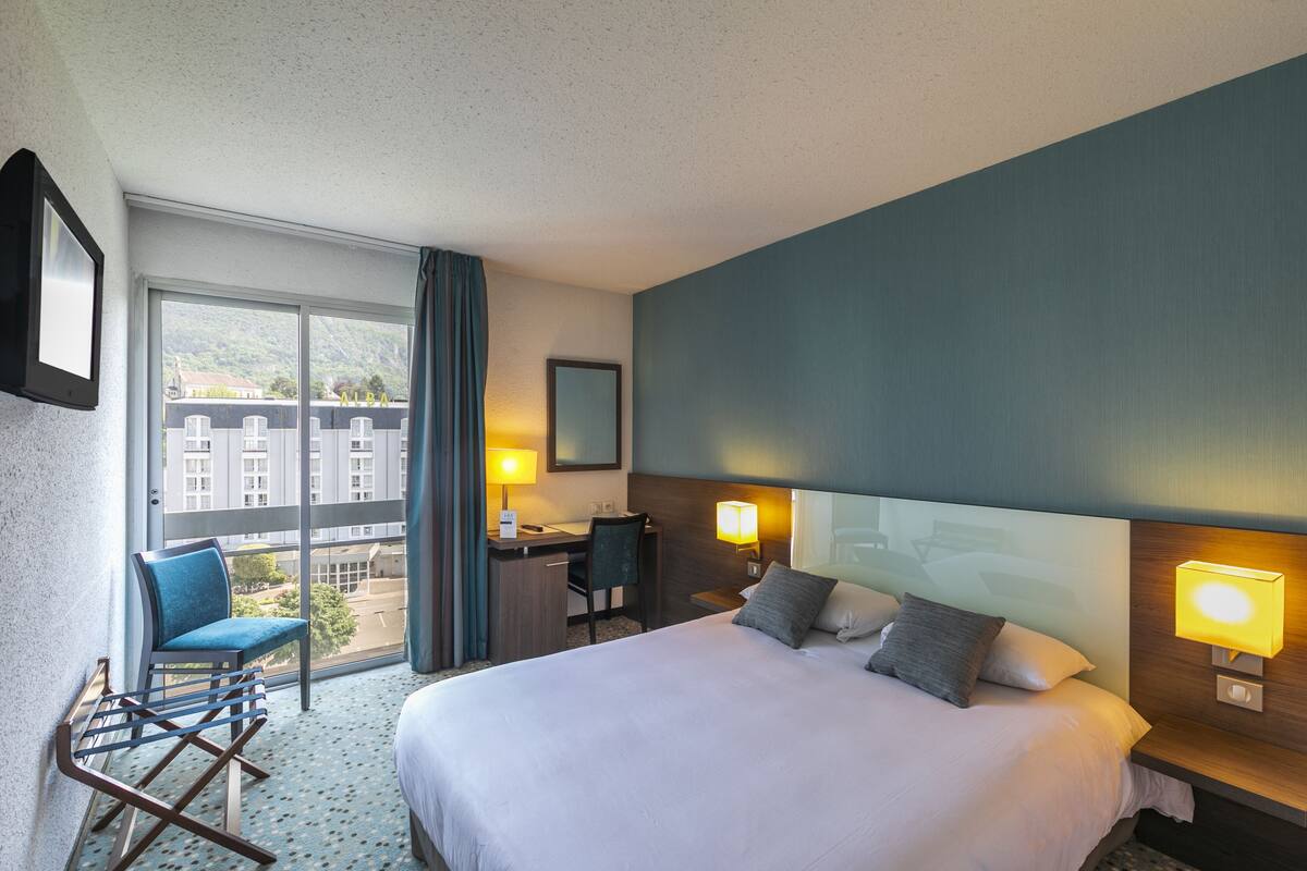 A hotel room with a large bed, a desk, a chair, and a view of buildings through the window.