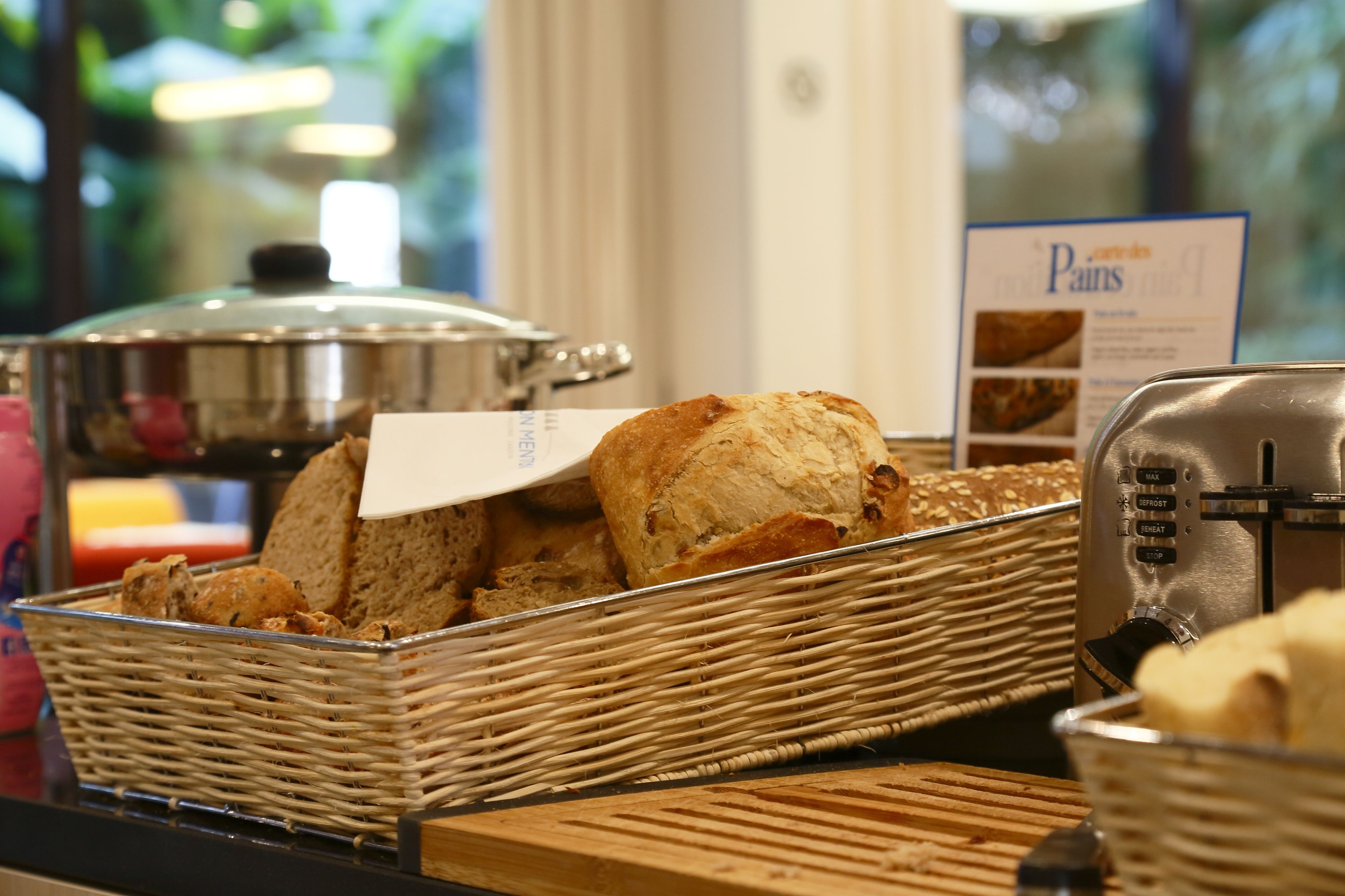 Un buffet de petit-déjeuner proposant diverses sortes de pains et de viennoiseries présentées dans des paniers en osier, une machine à café et un panneau indiquant le menu.