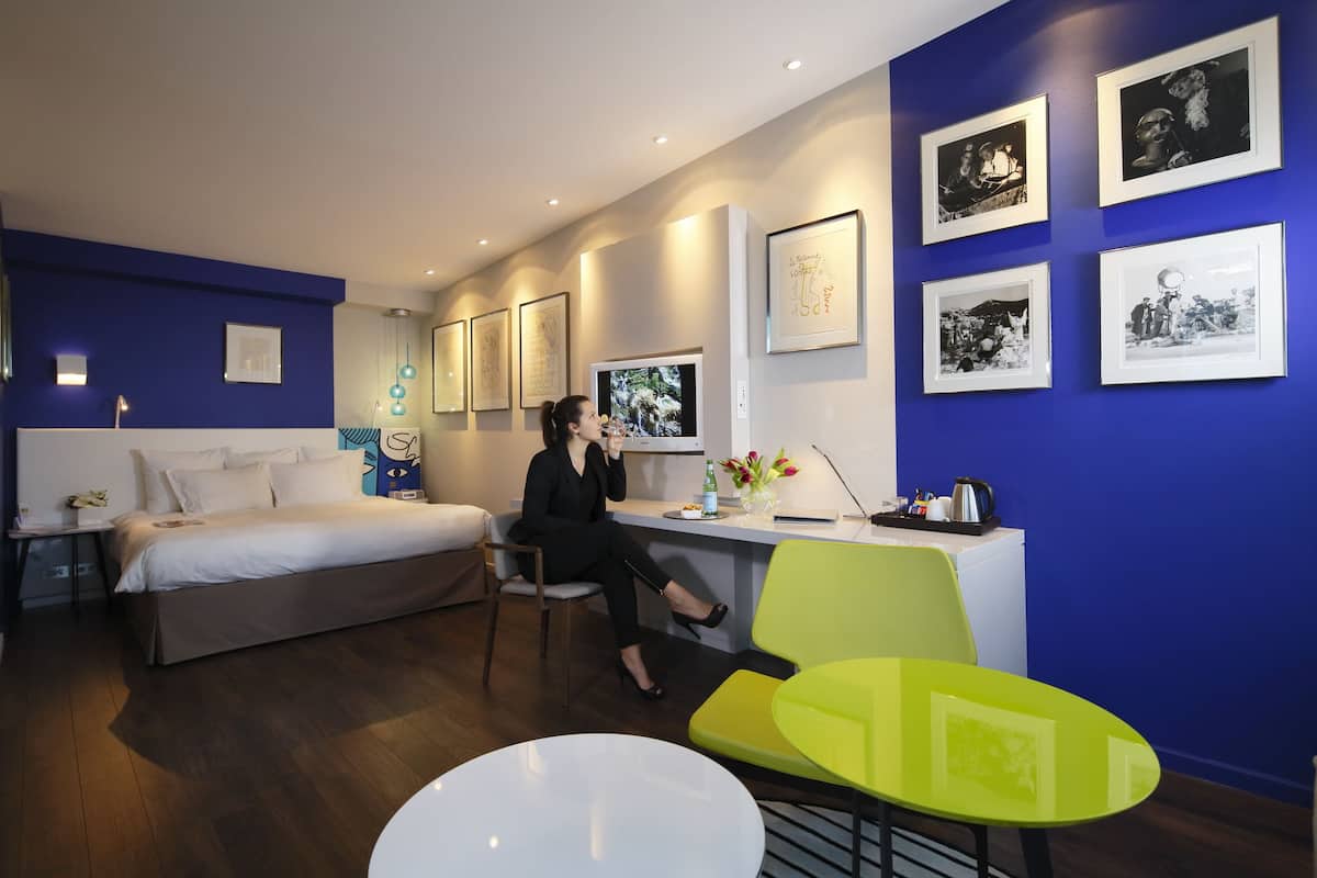 Une chambre d’hôtel moderne, équipée d’un lit, d’un bureau et de chaises. Des tableaux encadrés ornent le mur, et un téléviseur est posé sur le bureau.