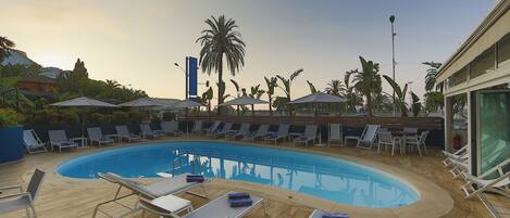 A pool area with lounge chairs and umbrellas, surrounded by palm trees and a wooden deck.