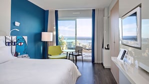 A hotel room with a bed, a desk with a computer, a chair, a lamp, and a large window with a view of the ocean. - Hotel Napoleon (Menton)