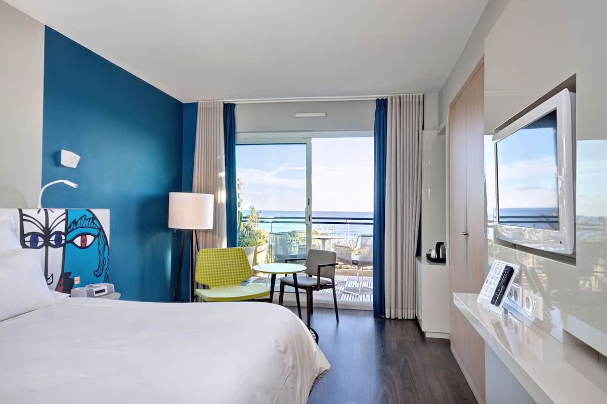 Une chambre d’hôtel avec un lit, un bureau équipé d’un ordinateur, une chaise, une lampe et une grande fenêtre donnant sur l’océan.