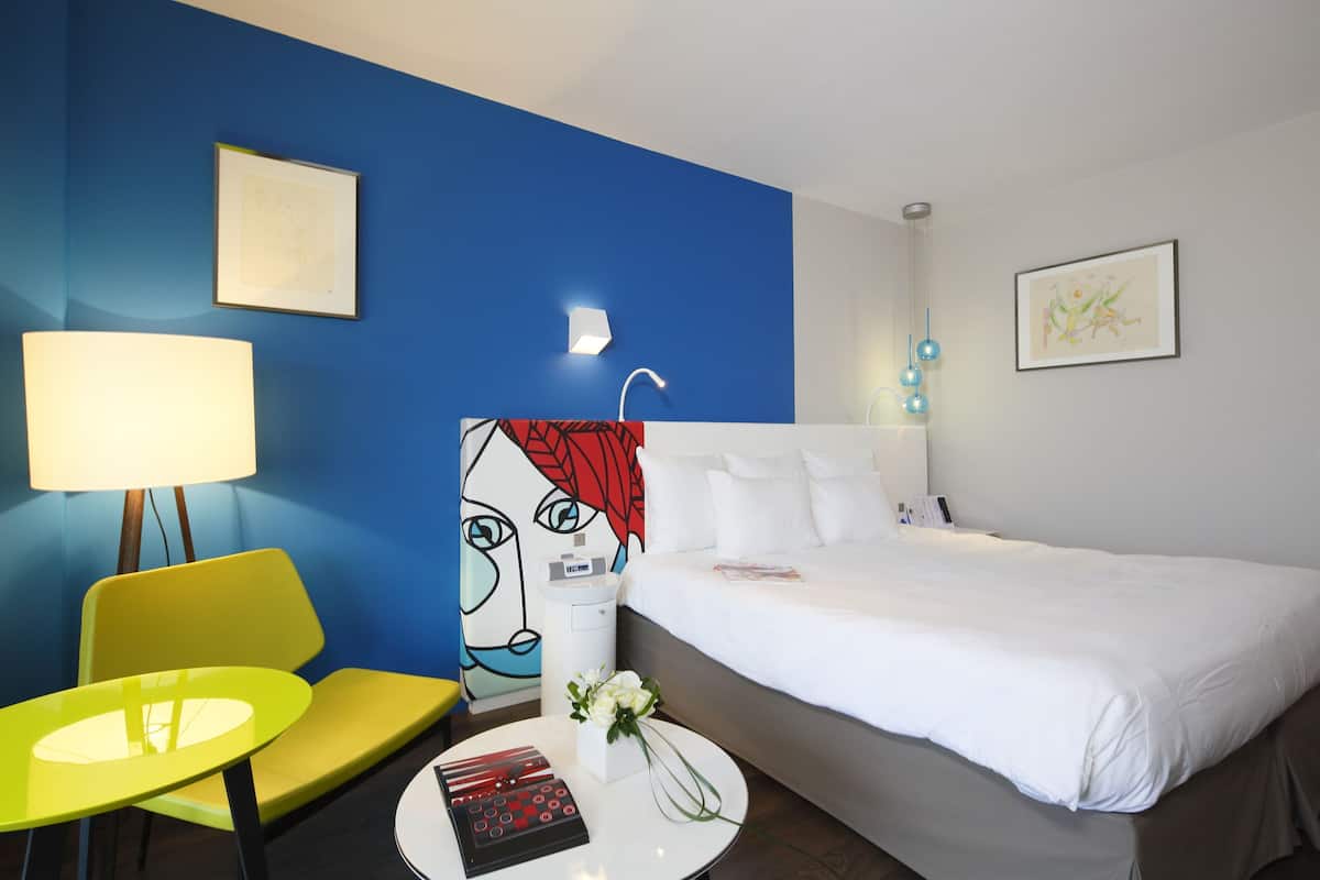Une chambre d’hôtel avec un lit, une chaise, une table, une lampe et des œuvres d’art accrochées aux murs.