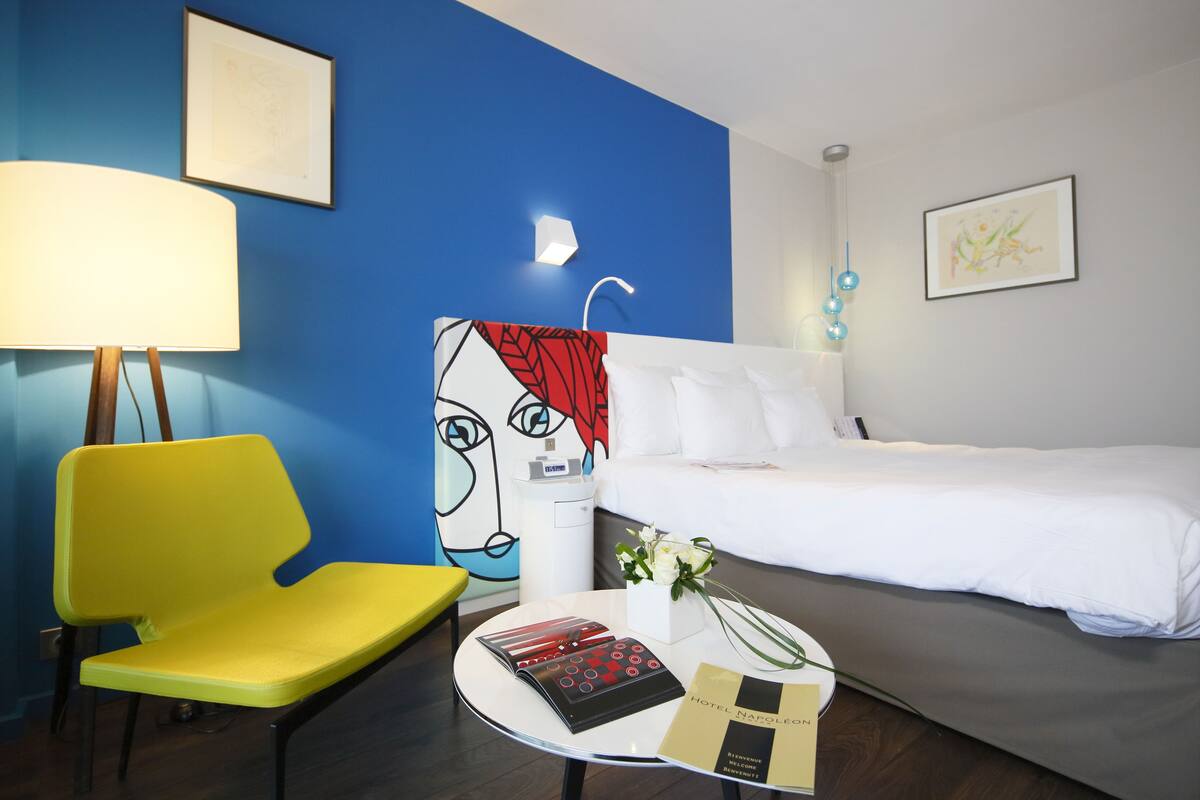 Une chambre d’hôtel avec un lit, une chaise jaune, une table avec un magazine, et un mur avec un détail bleu et un tableau encadré.