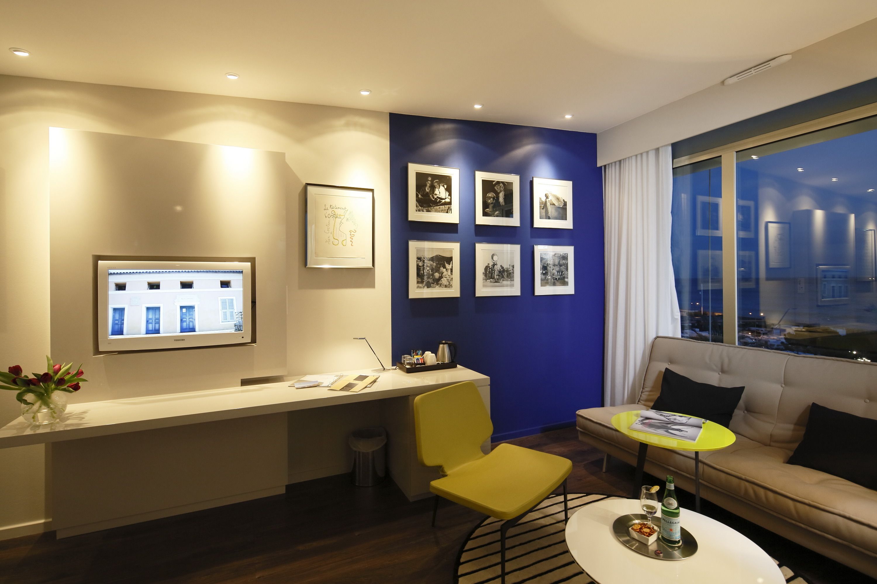 Habitación de hotel moderna con una recepción, un sofá, una pequeña mesa redonda y un gran ventanal con vistas a la ciudad.