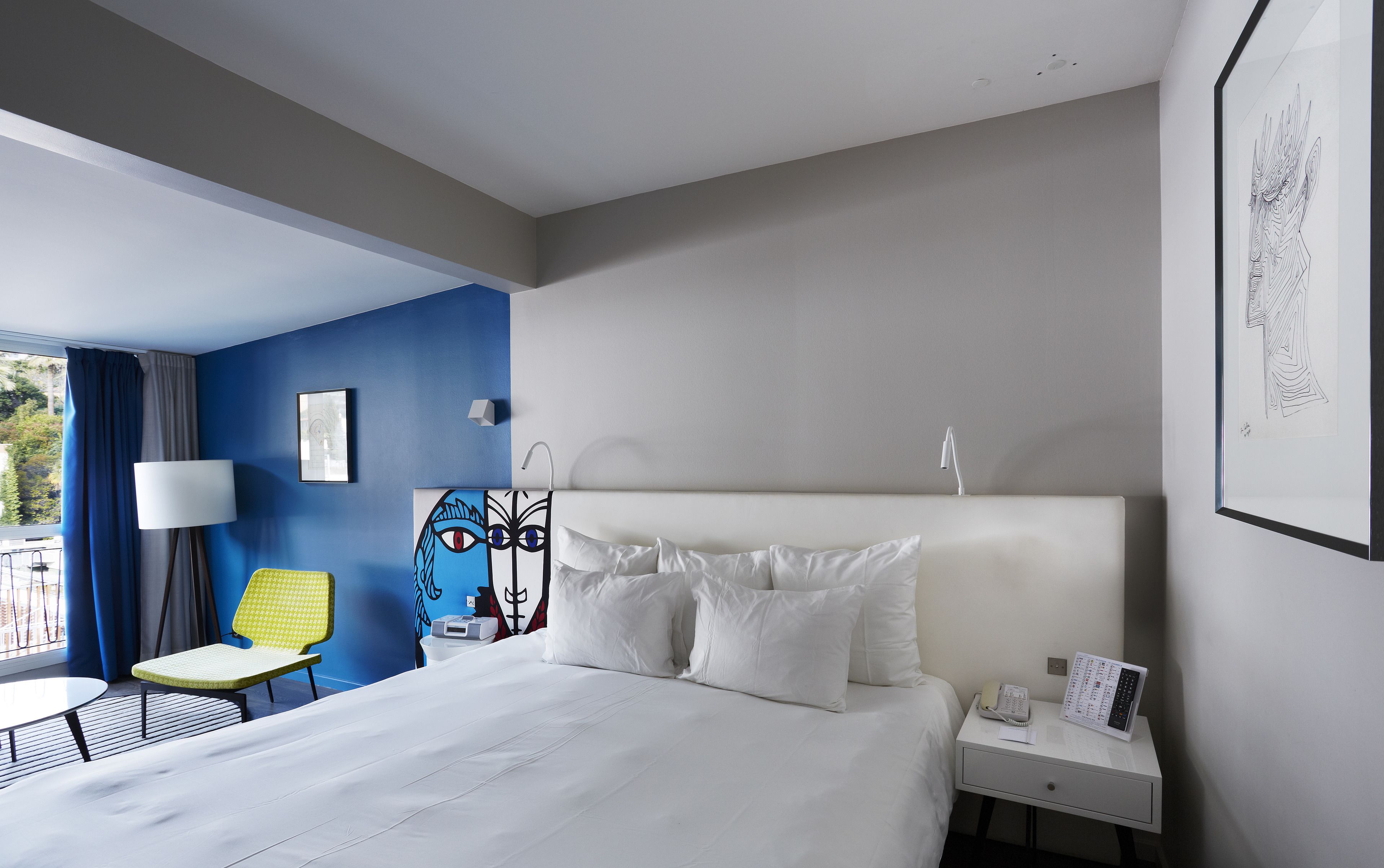 A hotel room with a bed, bedside table, lamp, chair, and artwork on the wall.