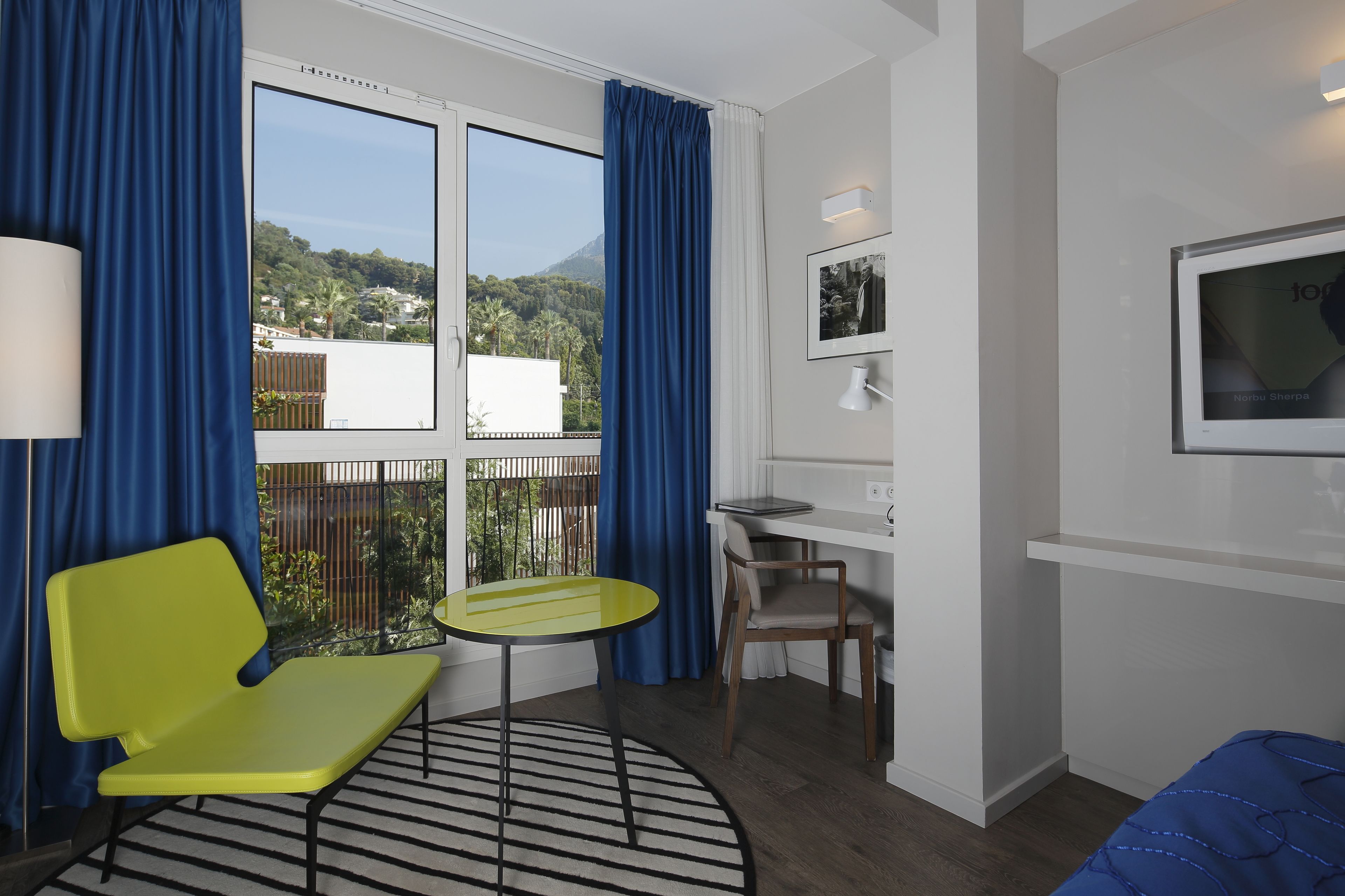 A room with a balcony, a yellow chair, a small round table, a desk, a chair, and a bed with a blue bedspread.