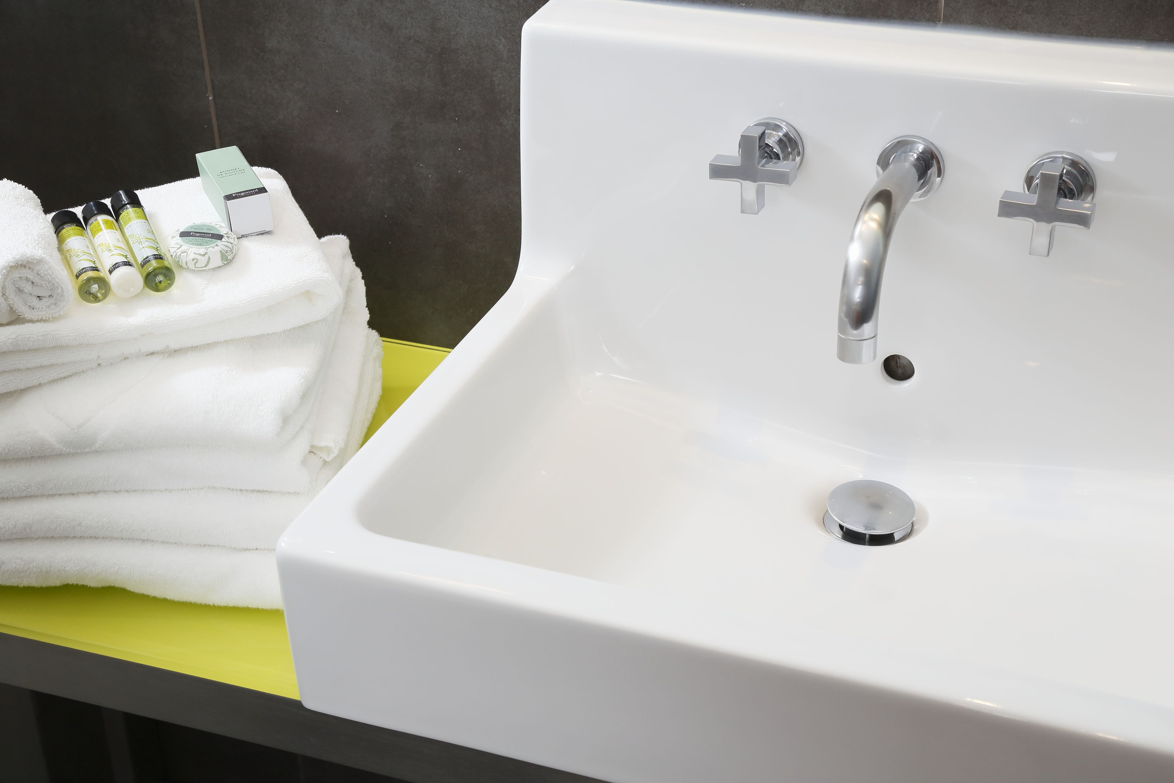 Un évier blanc avec deux robinets et un drain, une pile de serviettes blanches et quelques articles de toilette sur un comptoir jaune.