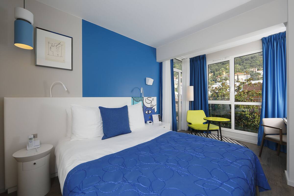 Une chambre d’hôtel avec un lit, une table de chevet, une chaise, une fenêtre avec des rideaux et une vue sur la verdure à l’extérieur.