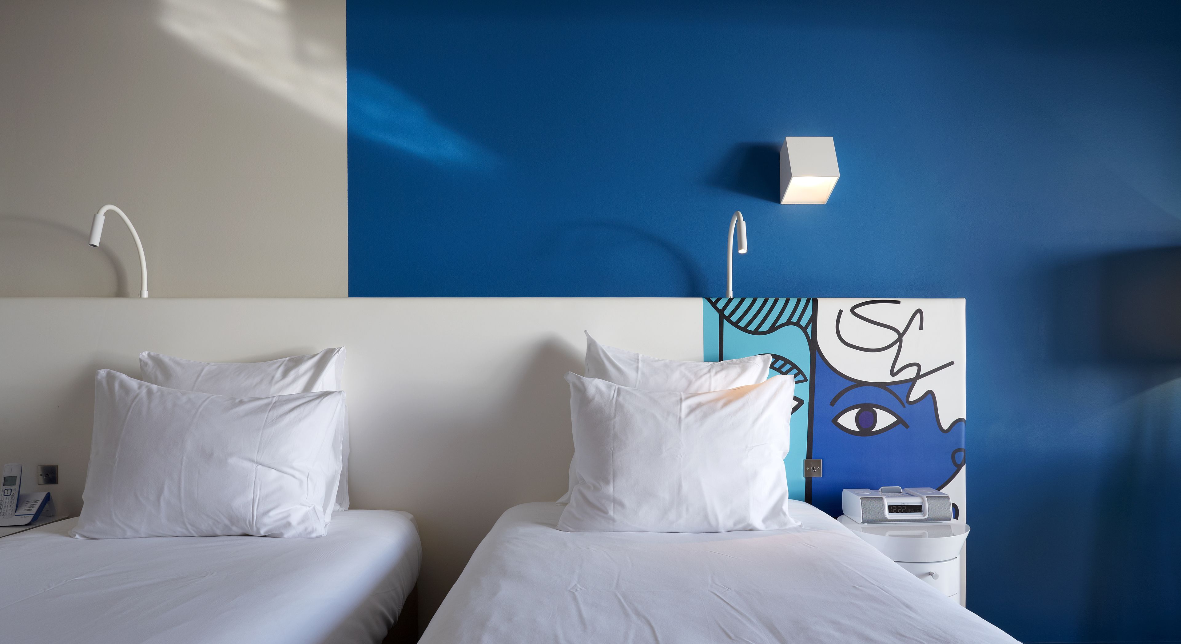 A hotel room with two beds, a bedside table with a lamp, a wall-mounted phone, and a blue wall with a graphic.