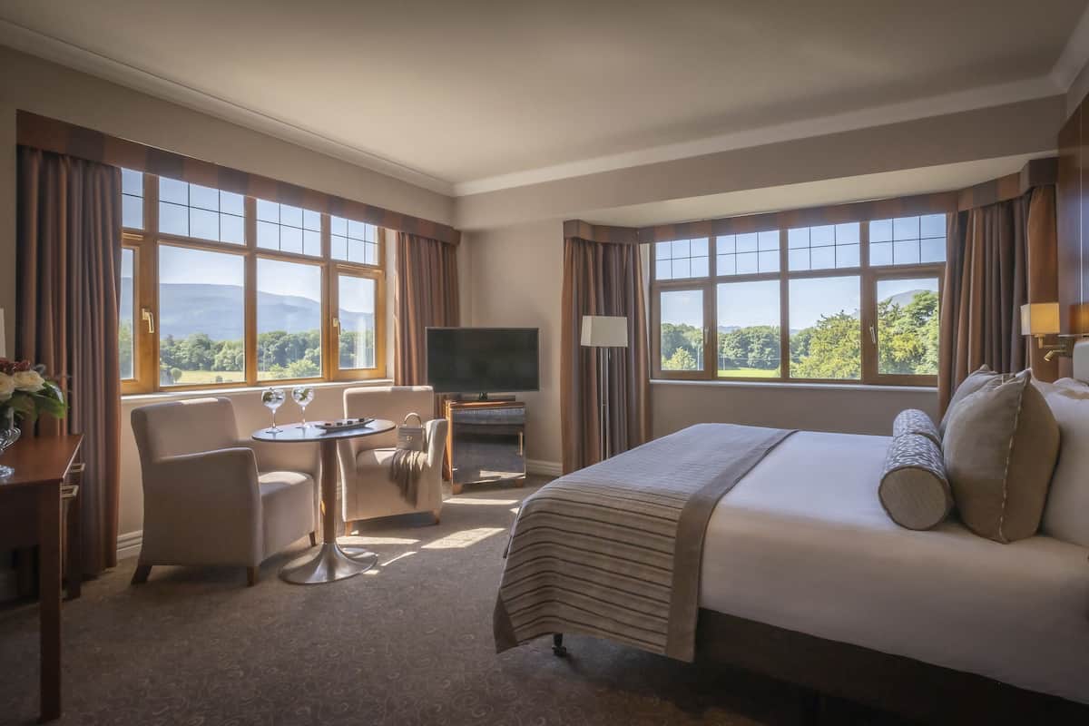 A hotel room with a bed, a desk with a chair, a TV, and large windows with curtains.