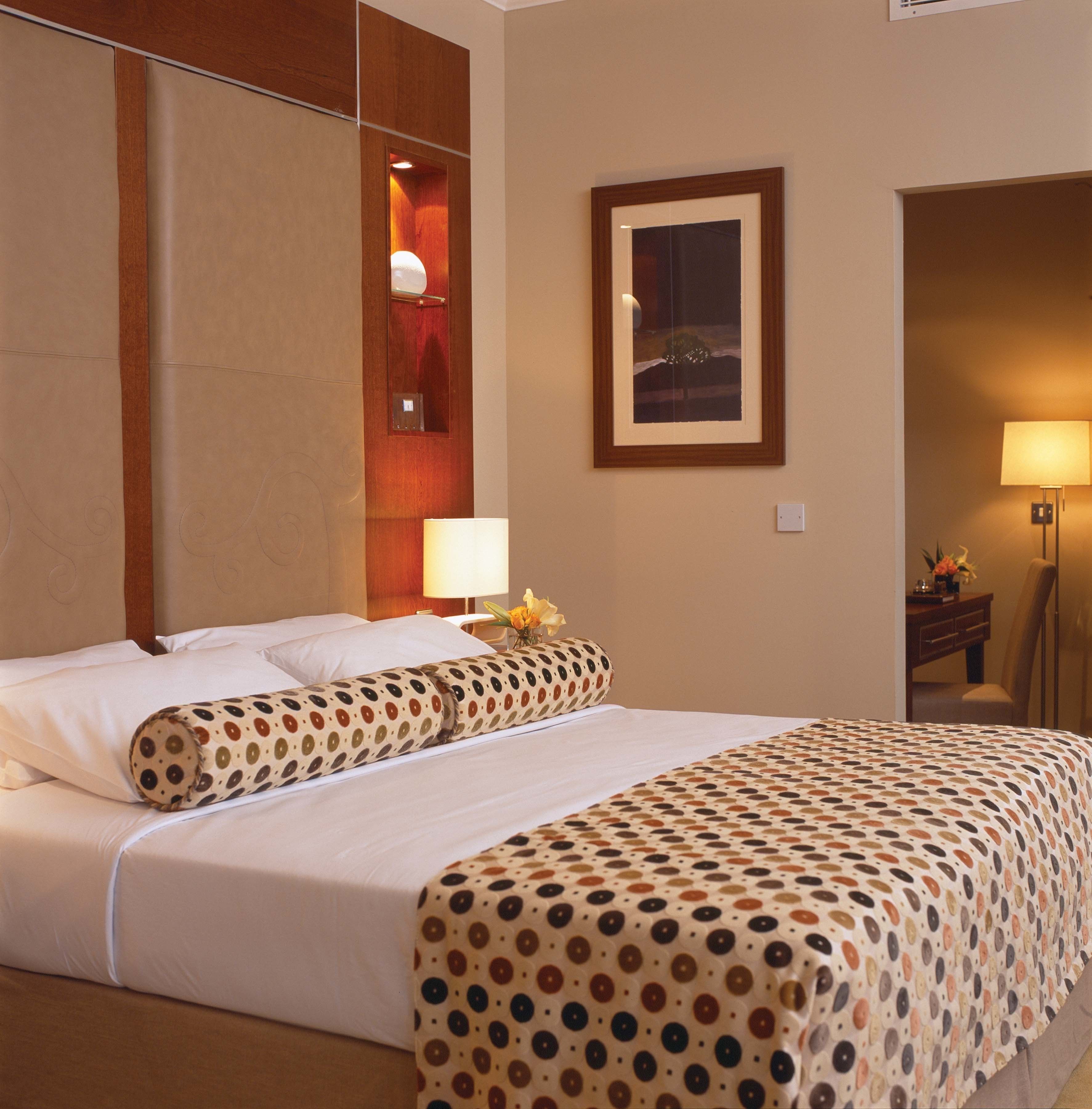 Une chambre d’hôtel avec un lit, une table de chevet avec une lampe, un tableau encadré au mur et une porte donnant sur une autre pièce.