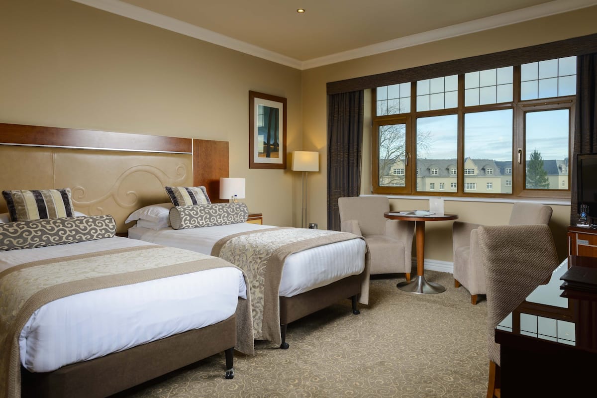 A hotel room with two beds, a desk, a chair, a lamp, and a window with a view.
