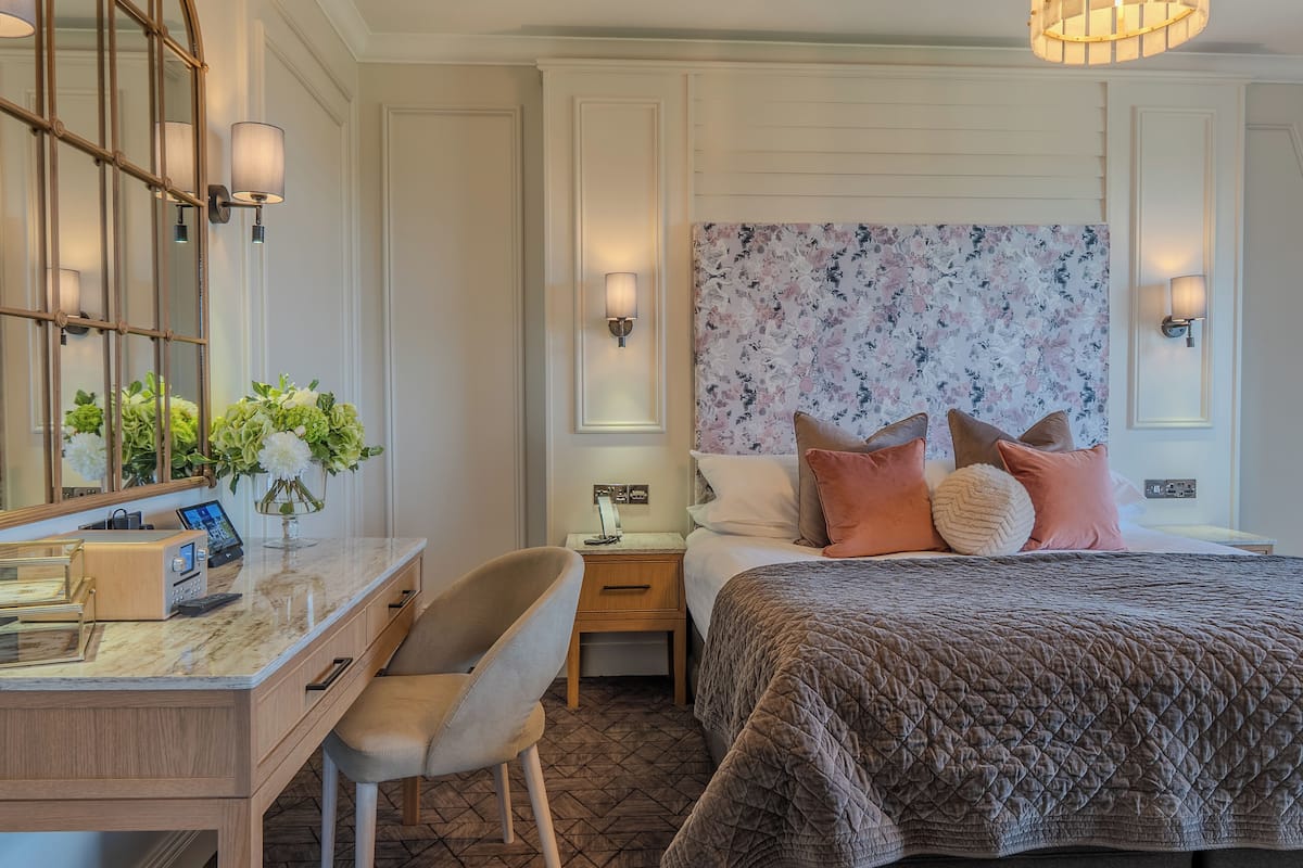 A hotel room with a bed, a desk with a chair, a nightstand, and a wall-mounted lamp.