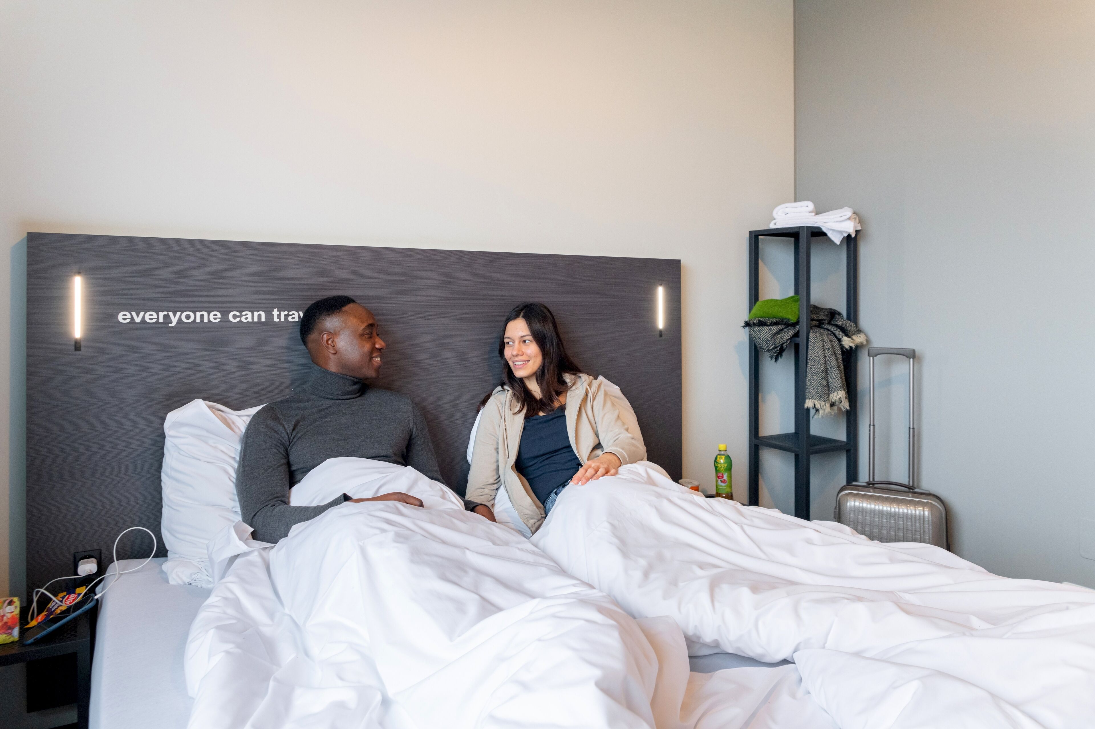 A hotel room with a bed, a nightstand, a suitcase, and a shelf with towels and a green pillow.