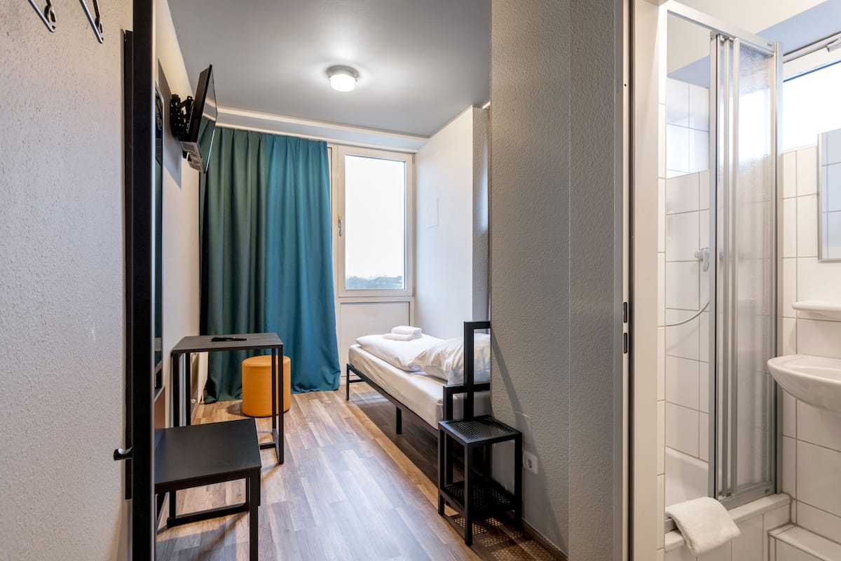 Ein kleines Zimmer mit Bett, Tisch, Stuhl, einem Fenster mit Vorhängen sowie einer Dusche.
