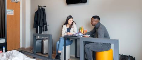 A hotel room with a bed, a desk, a TV mounted on the wall, and a suitcase on the floor.