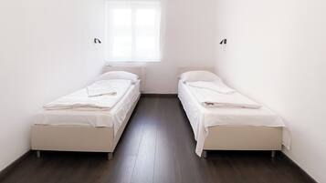Ein schmales Zimmer mit zwei Einzelbetten, weißer Bettwäsche und einem Fenster mit leichtem Vorhang.