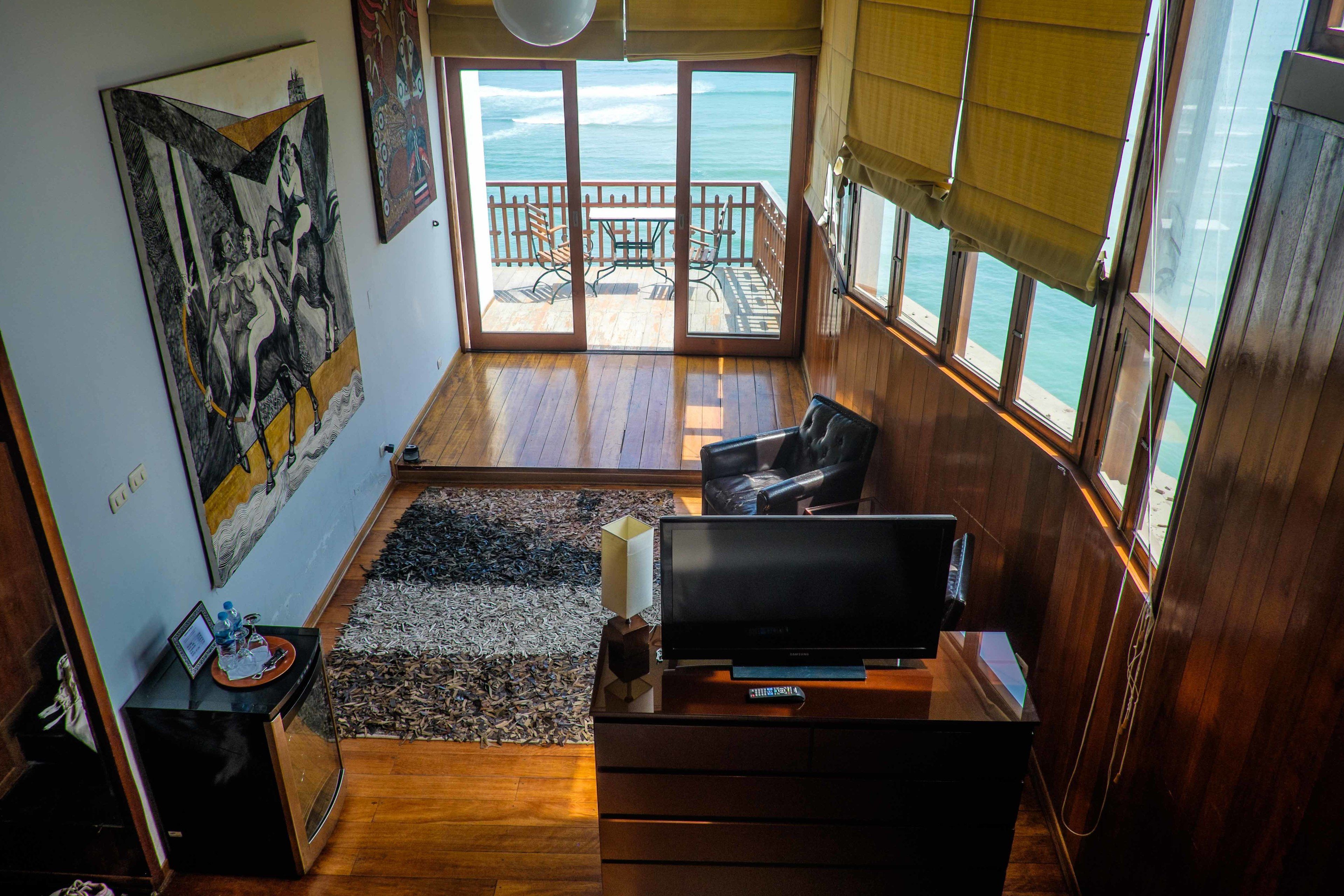 Una habitación con una televisión grande, piso de madera, un cuadro en la pared y vista al océano a través de amplias ventanas.