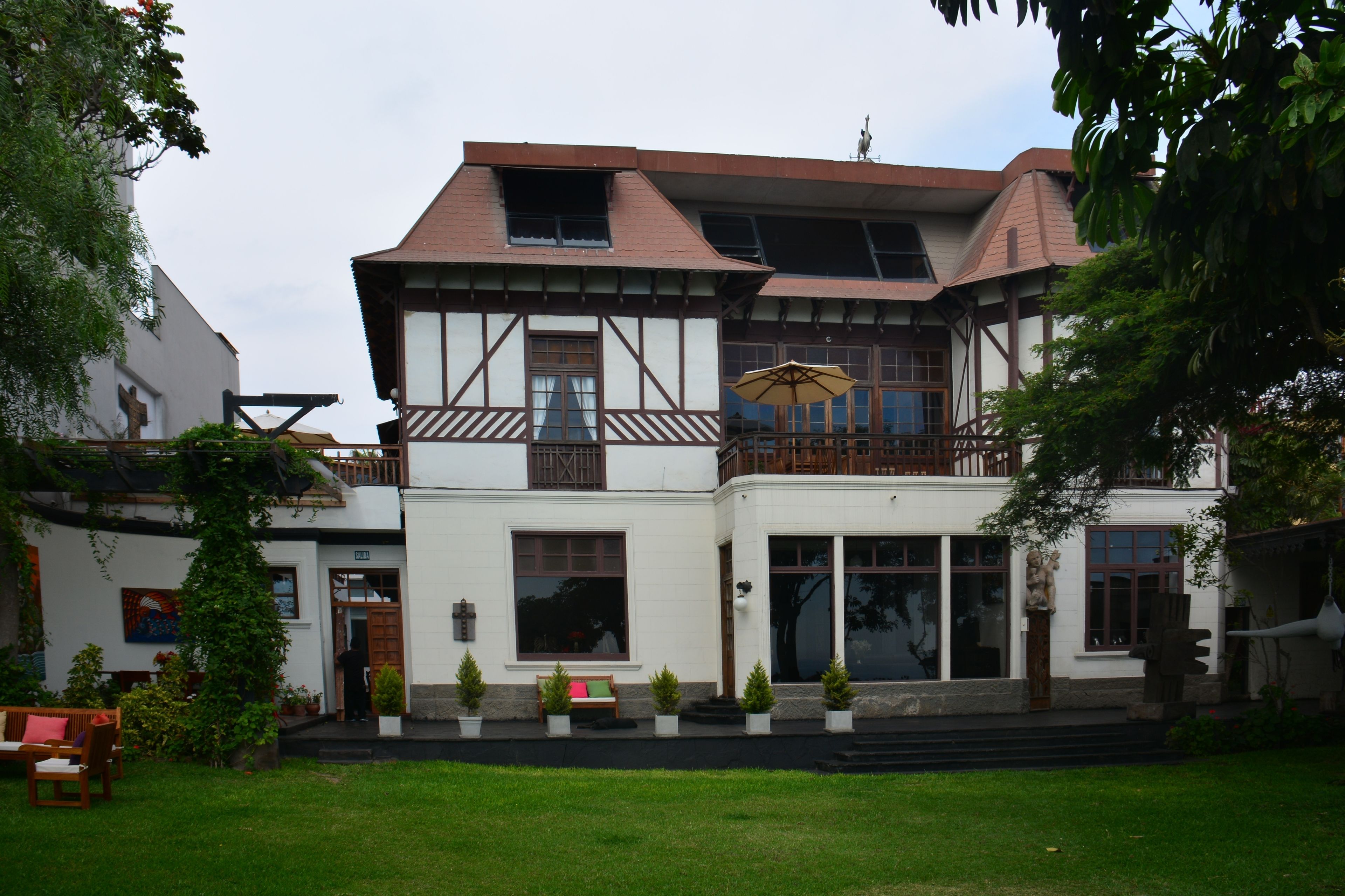 Una casa tradicional con entramado de madera, con un balcón, una puerta principal y un pequeño jardín con un banco y macetas.