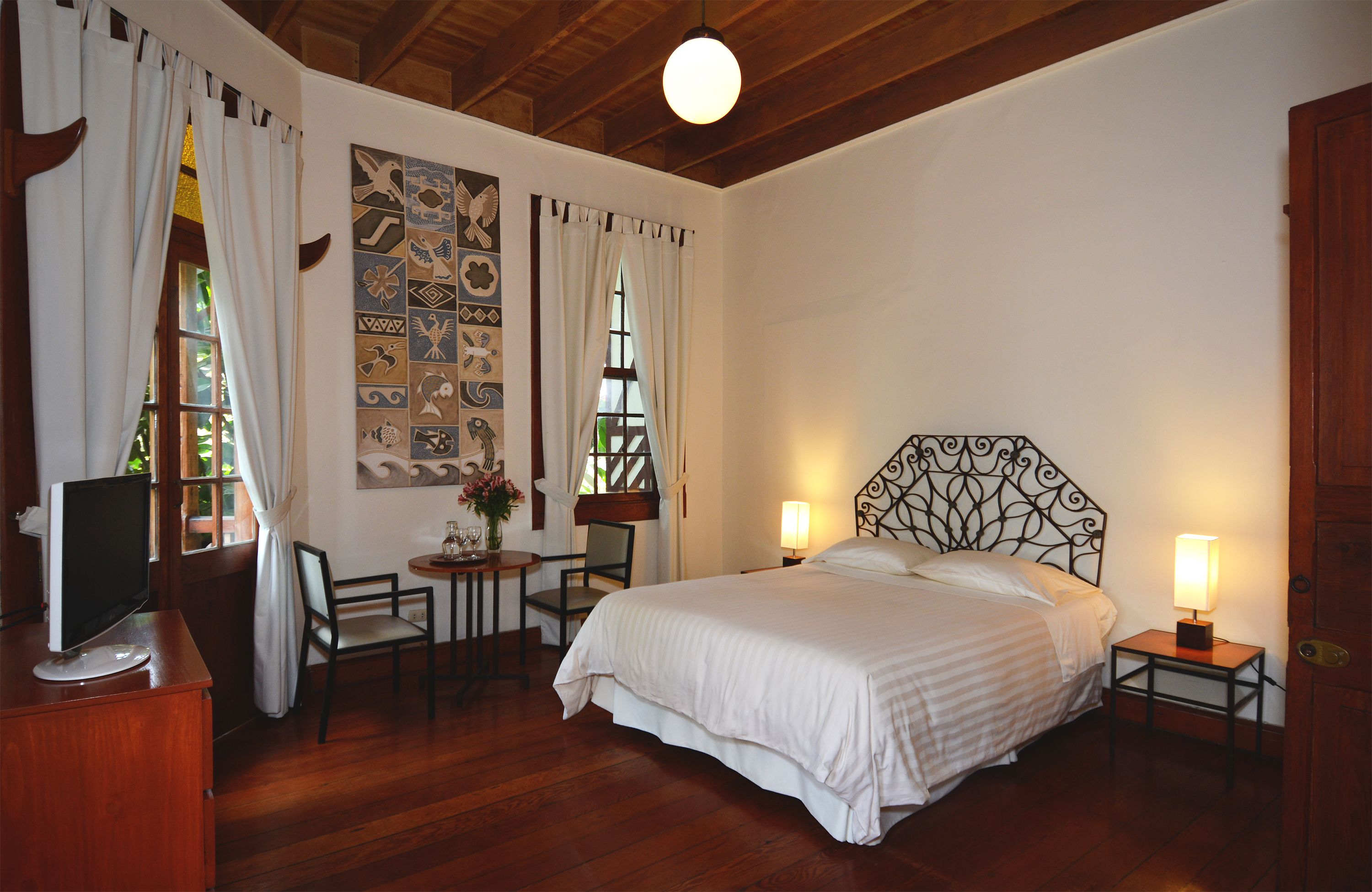 A bedroom with a large bed, two bedside tables with lamps, a desk with a chair, a TV on a wooden cabinet, and a wall with decorative tiles.