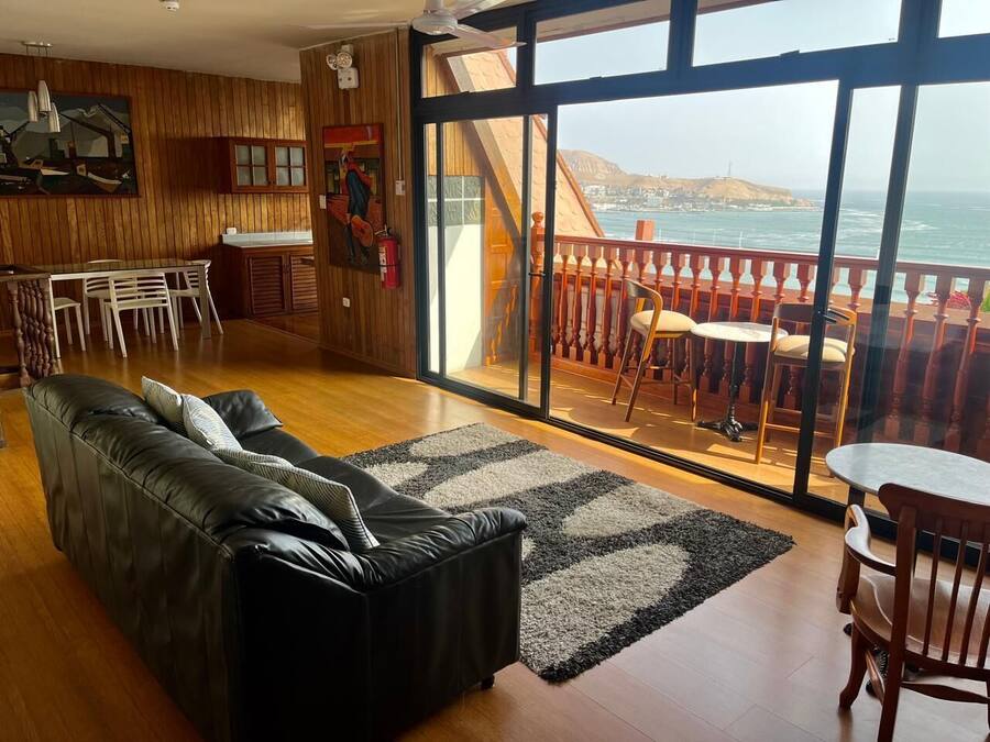 Un salón con un sofá de cuero negro, un comedor con mesa y sillas, y un amplio ventanal corredero que da a un balcón con vistas al océano.