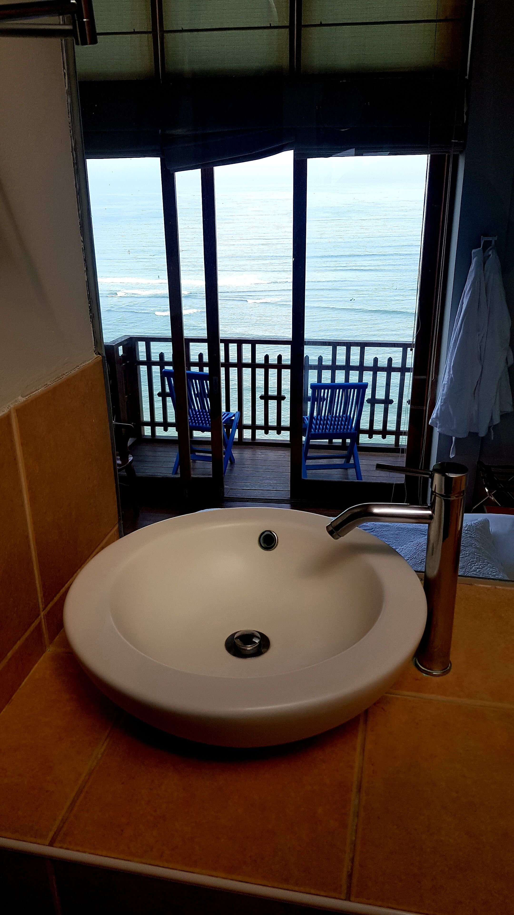 Une salle de bain avec un grand lavabo, une vue sur l’océan par la fenêtre et des chaises bleues à l’extérieur.