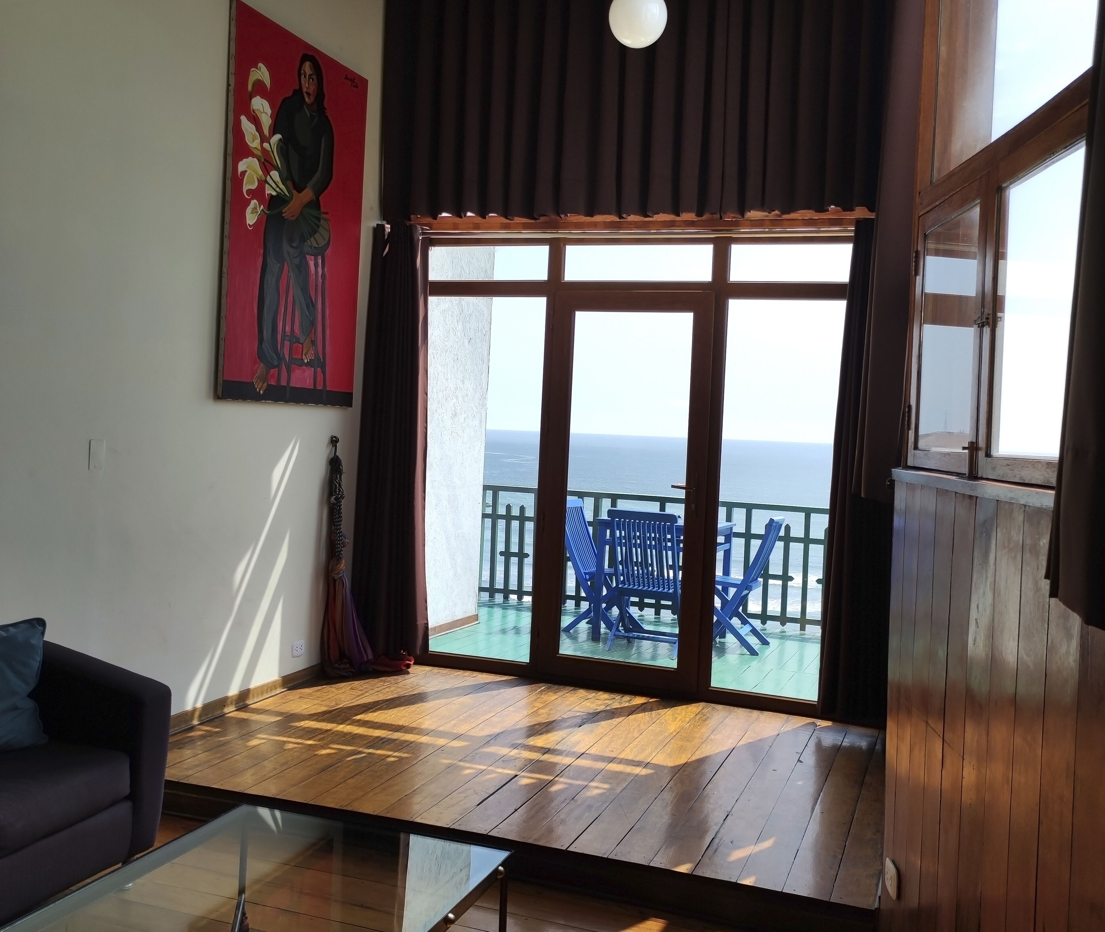 Une pièce avec un plancher en bois, une table basse en verre, un canapé foncé, un tableau accroché au mur et une vue sur la mer par une grande fenêtre.