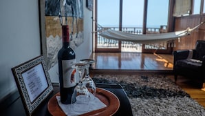 Una habitación con un ventanal grande que da a una playa, una hamaca, un menú enmarcado, una botella de vino y copas sobre una bandeja.