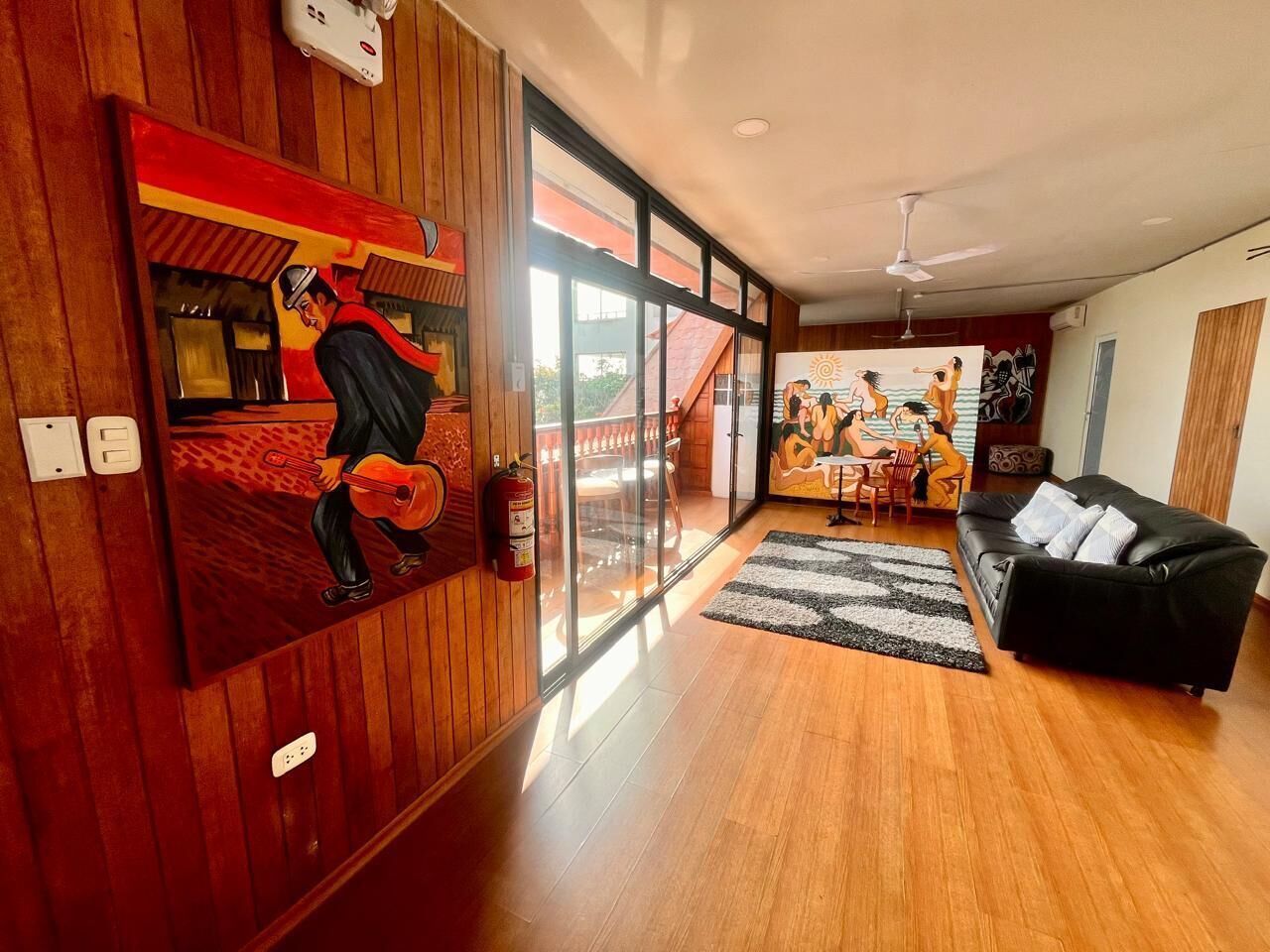 A modern living room with a wooden floor, a black sofa, a glass door leading to a balcony, and a painting of a man playing a guitar.