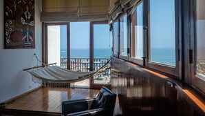 Una habitación con hamaca, suelo de madera, un ventanal con vista al océano y un cuadro en la pared.
