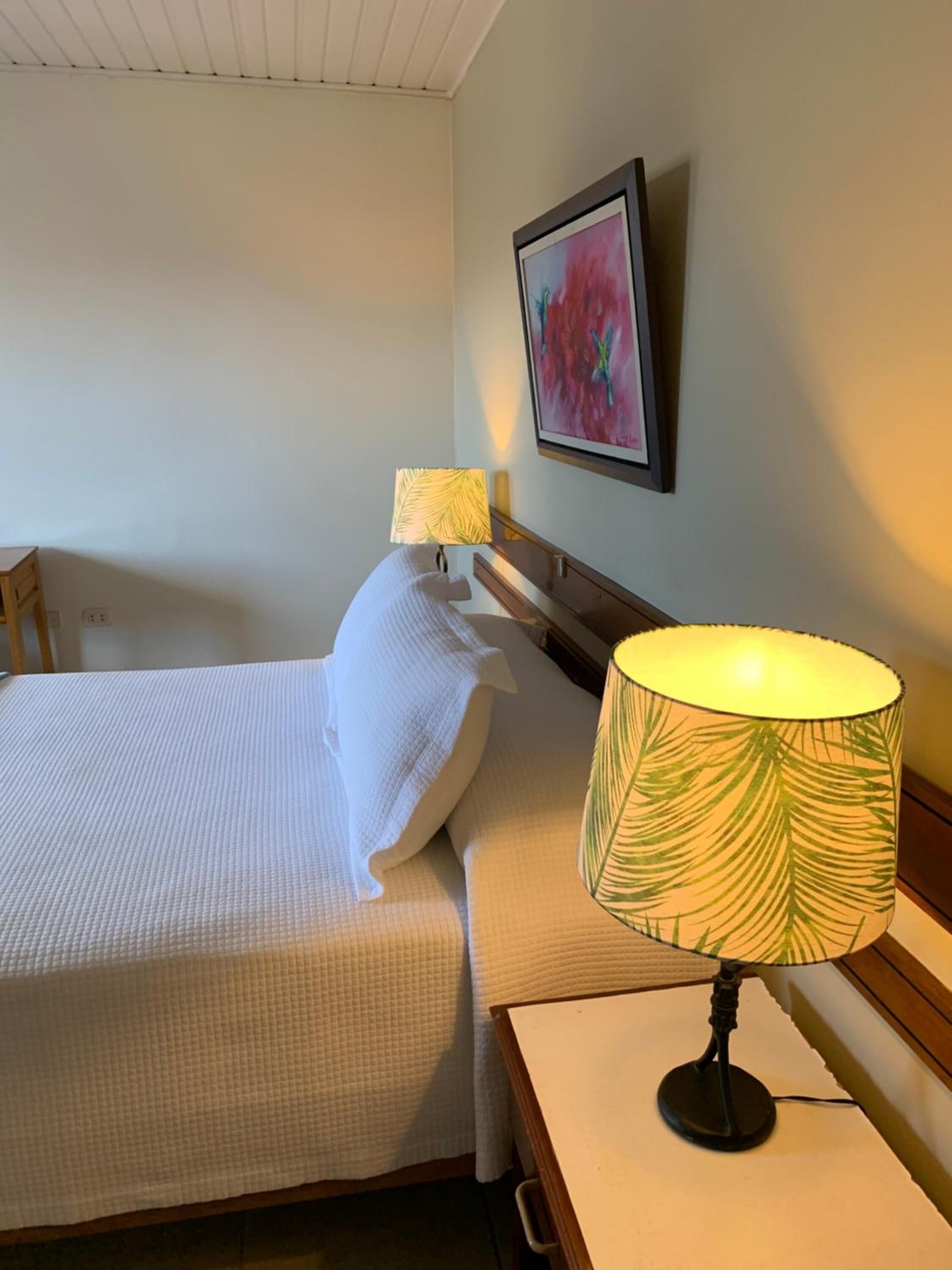 A hotel room with a bed, a nightstand, a lamp, and a framed picture on the wall.