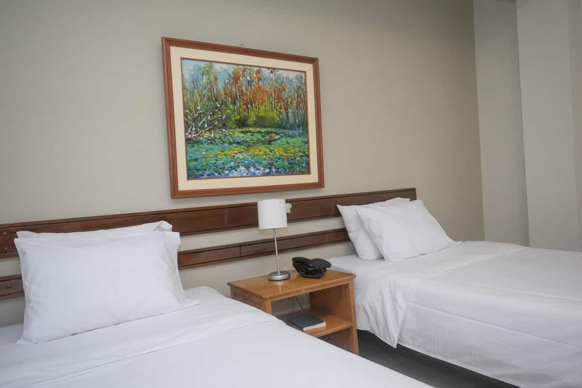 Une chambre d’hôtel avec deux lits simples, une table de chevet avec une lampe, un téléphone et un tableau encadré au mur.