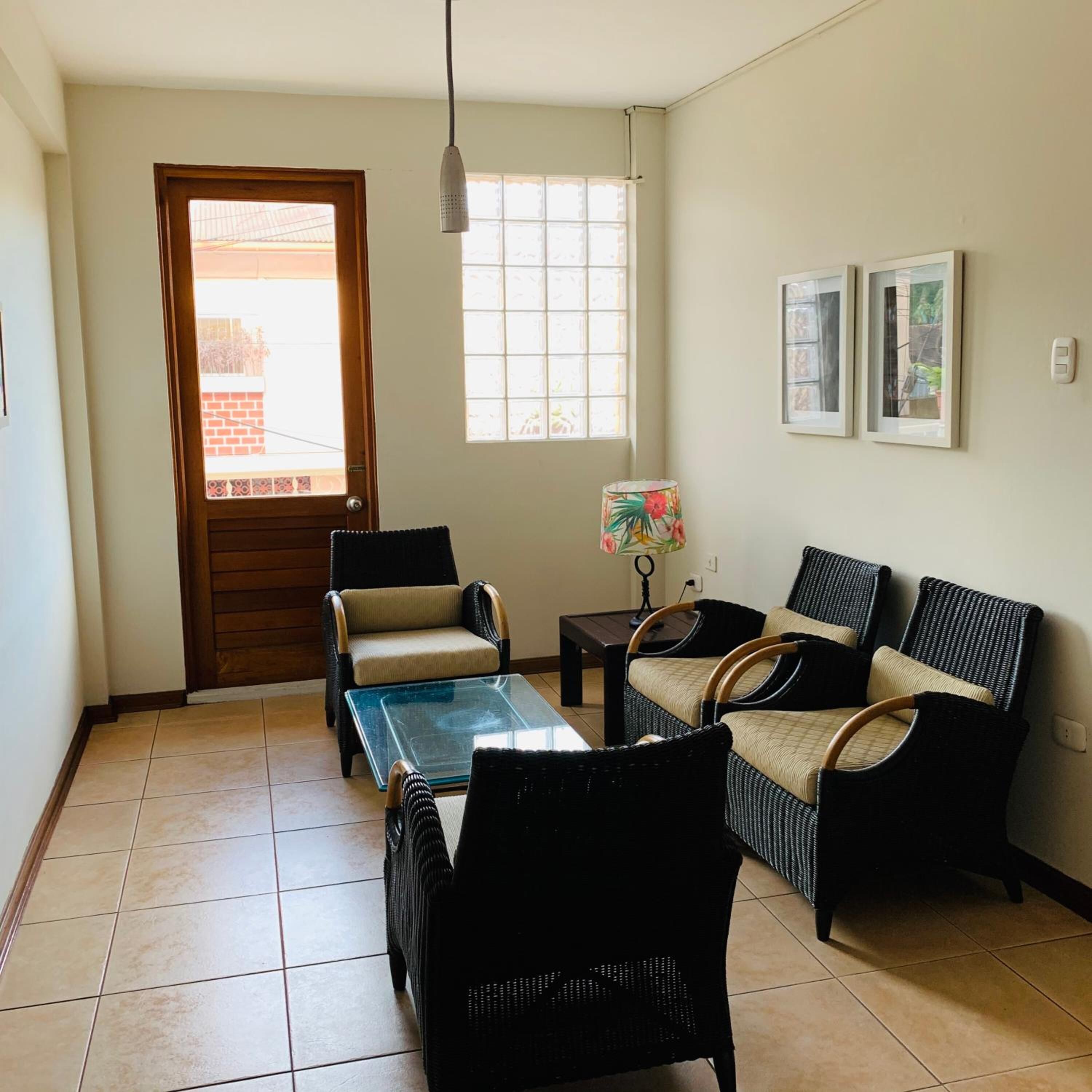 Una sala de estar con una mesa de centro de vidrio, dos sofás de mimbre, una silla, una lámpara y una puerta que da al exterior.