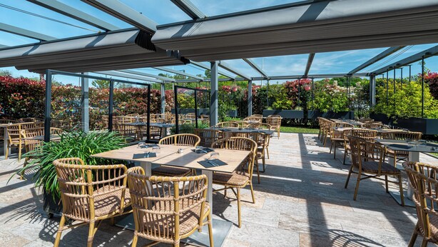An outdoor patio with tables and wicker chairs, surrounded by glass walls and a view of greenery. - Hôtel Loreak (Bayonne)