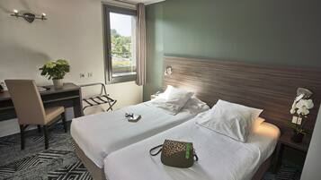 Une chambre d’hôtel avec deux lits, un bureau, une chaise, une plante en pot et une fenêtre donnant sur un paysage verdoyant.