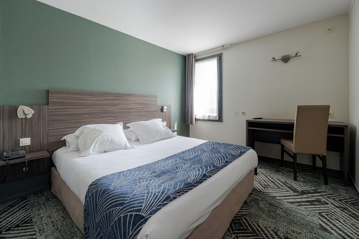 A hotel room with a bed, desk, chair, and a telephone on the nightstand.