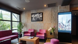 A modern hotel lobby with red upholstered seating, a wooden coffee table, a large window, a framed photograph, and a digital screen displaying a cityscape. - Hôtel Loreak (Bayonne)