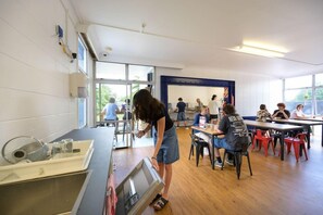 A classroom with tables and chairs, a person standing by a counter, and a window providing natural light. - Base Rotorua - Hostel (Rotorua)