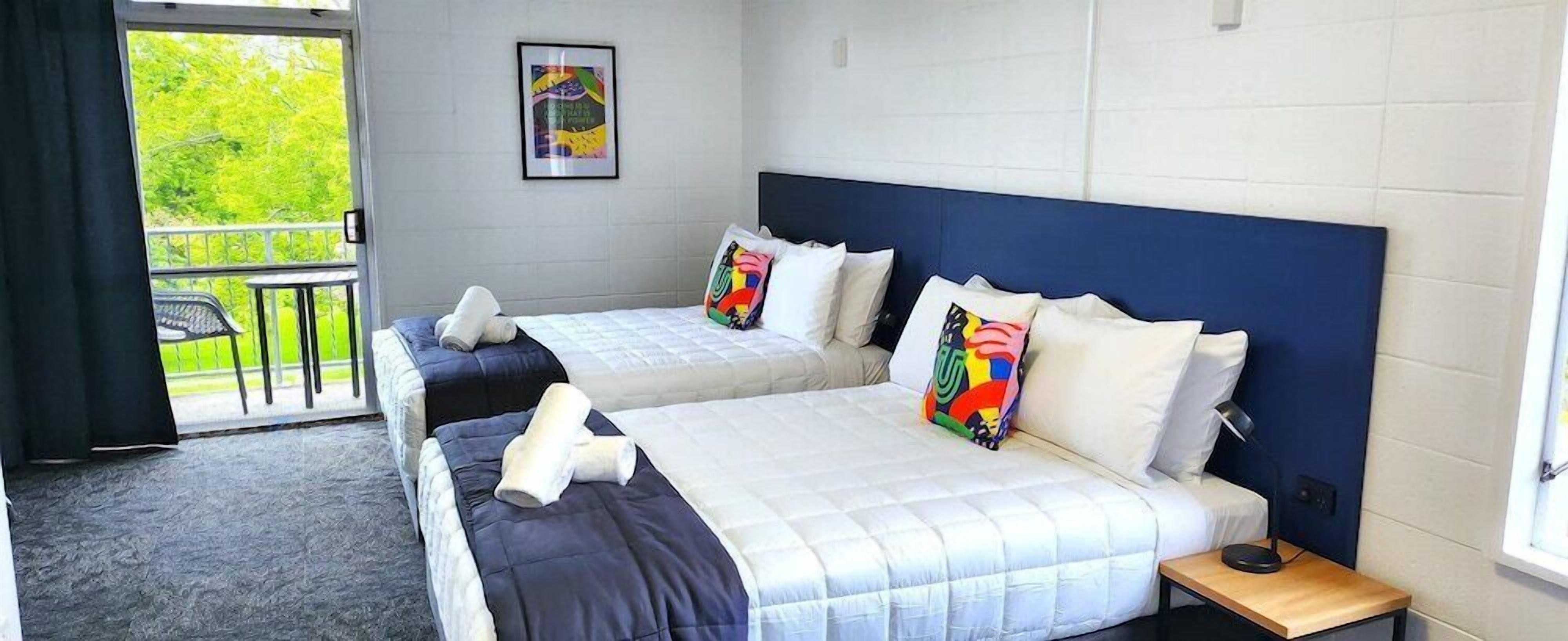A hotel room with two beds, a balcony, and a view of greenery.