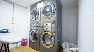 A laundry area with two front-loading washing machines, a drying machine, an ironing board, a trash bin, and a laundry basket.