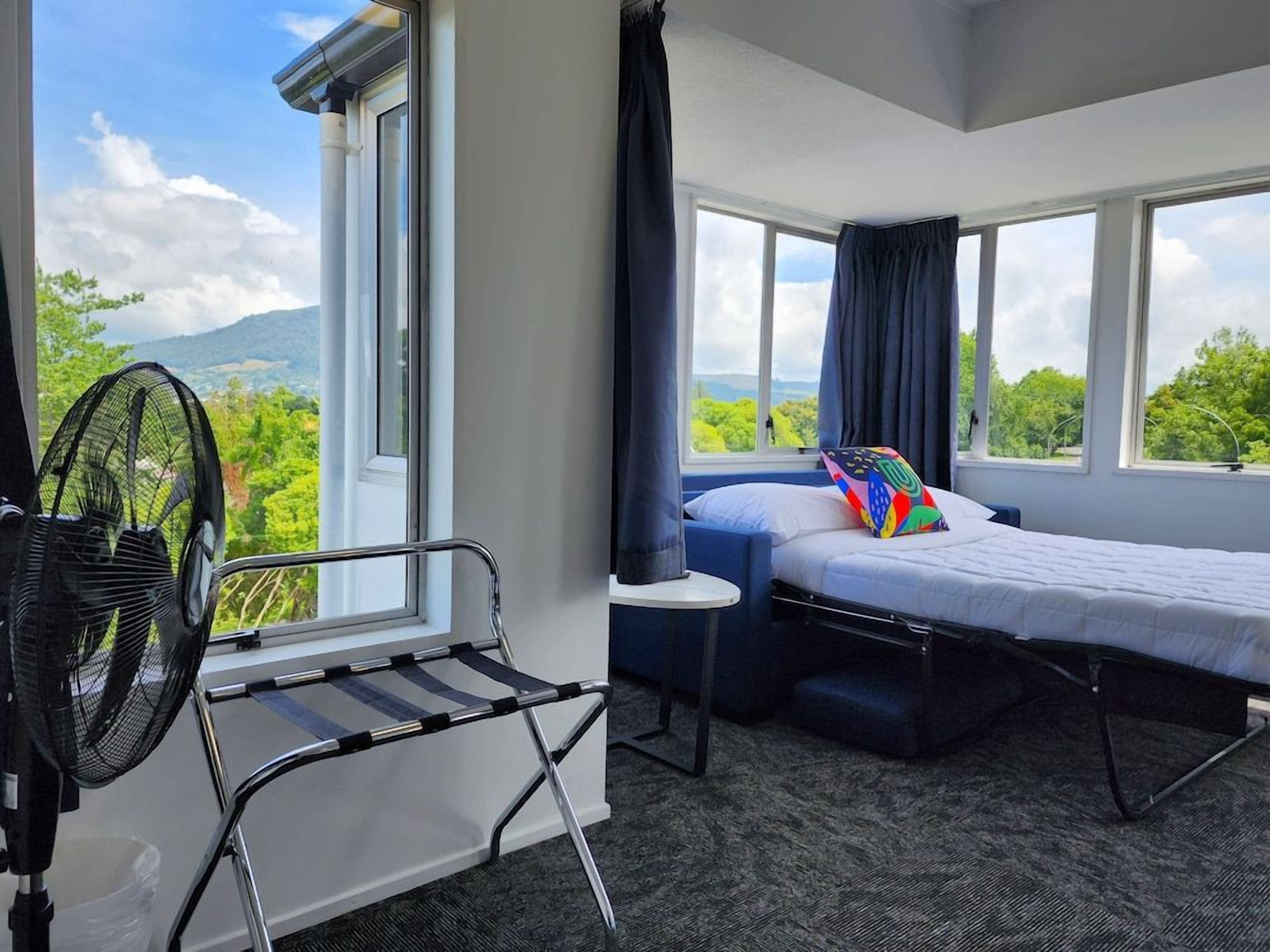 A room with a bed, a chair, a standing fan, and a window with a view of greenery.