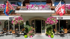 Entrada de hotel con balcón, mobiliario exterior y arreglos florales colgantes.