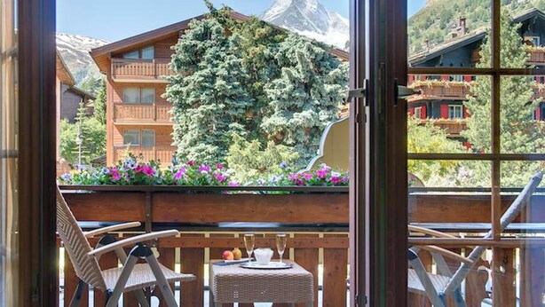 Un balcón con sillas de mimbre y una mesa, con vistas a una montaña donde se distingue una construcción de estilo chalet y un árbol en primer plano.