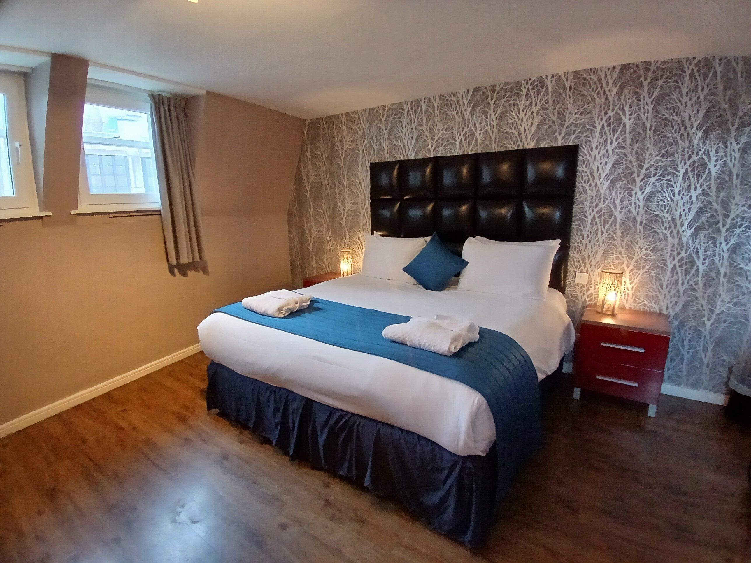A neatly made bed with a blue bedspread and white linens, a dark headboard, and a nightstand with a lamp.