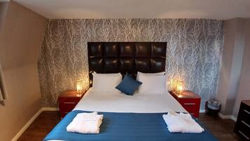A neatly made bed with a blue bedspread and white linens, flanked by two bedside tables with lamps, in a room with patterned wallpaper and a dark headboard.