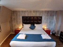 A neatly made bed with a blue bedspread and white linens, flanked by two bedside tables with lamps, in a room with patterned wallpaper and a dark headboard.