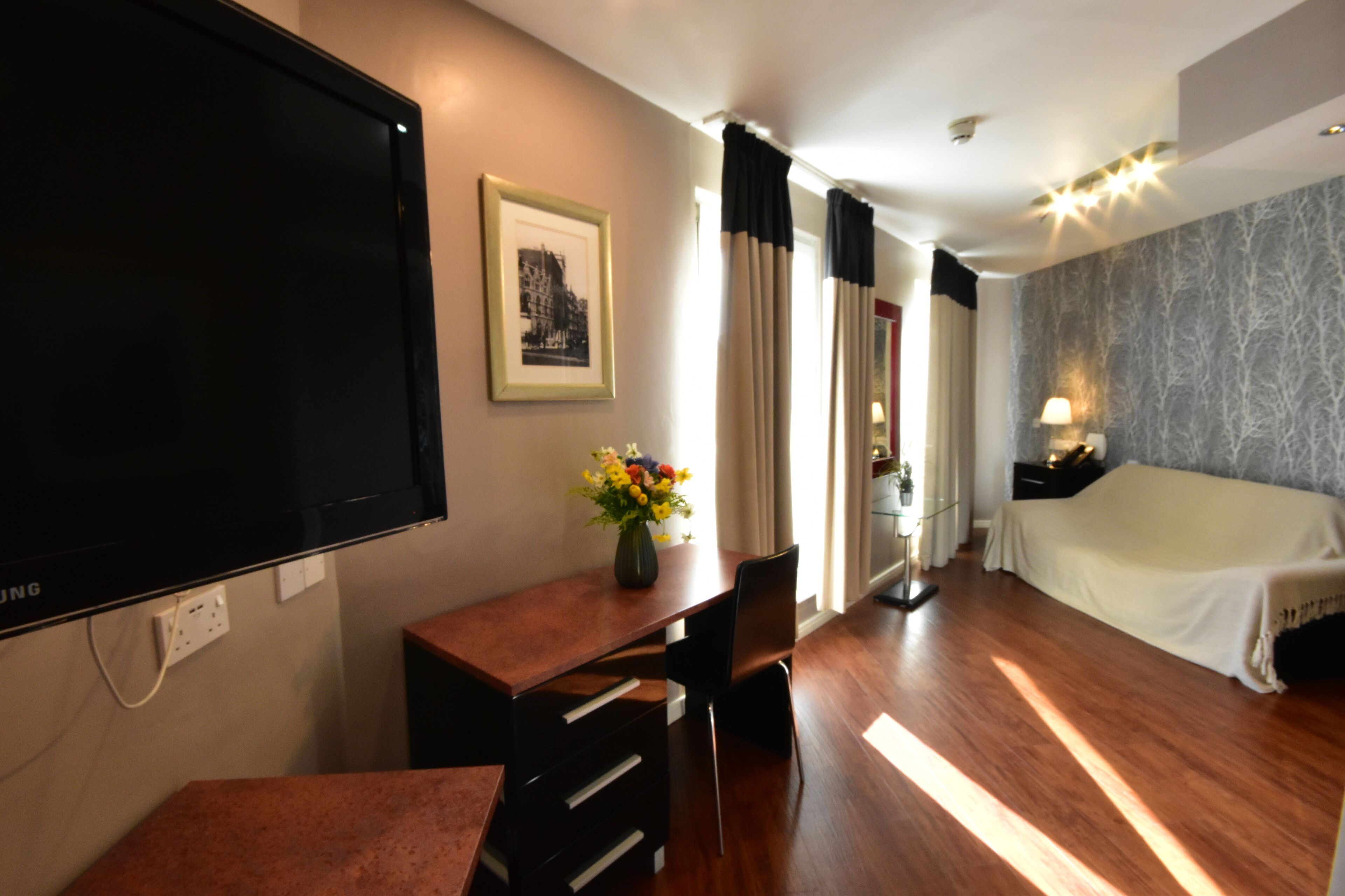 A hotel room with a bed, a desk with a chair, a TV, a framed picture, and a vase with flowers.